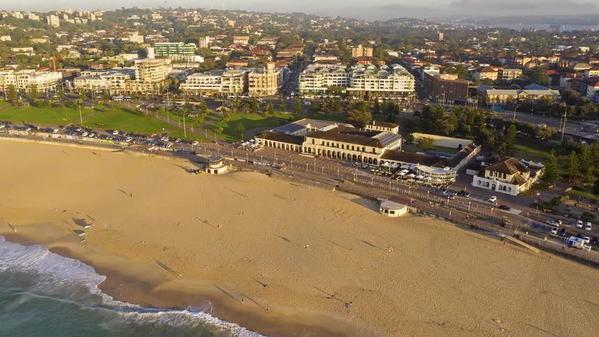 An aerial pull-back shot reveals the iconic Bondi Pavilion, the golden sand, and gentle waves of Sydney's Bondi Beach at sunrise