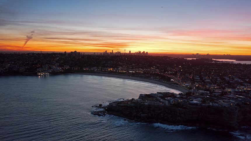 An aerial time-lapse of a beautiful coastal city as a vibrant sunset fades into twilight, with the distant city skyline silhouetted against the colorful sky