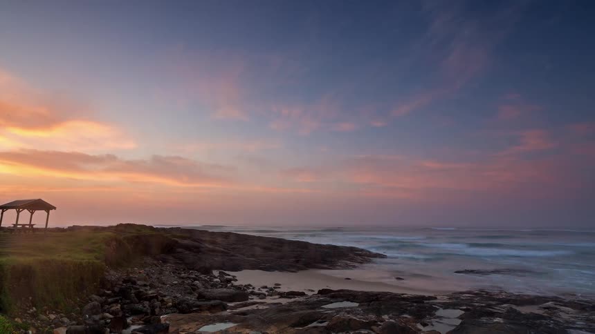 A serene timelapse captures the changing colors of a beautiful sunrise over a rocky coastline, with mist rolling over the waves
