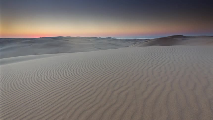 A serene time-lapse captures a vibrant sunset fading over rippled sand dunes, followed by rolling fog and a brilliant sunrise