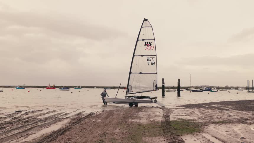 A sailor wades through muddy waters to launch a small dinghy into the harbor on an overcast day