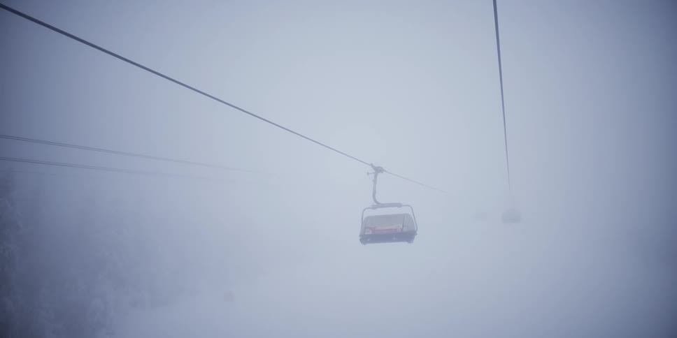 A ski lift and empty chairlifts move slowly through the dense fog on a snowy mountain slope