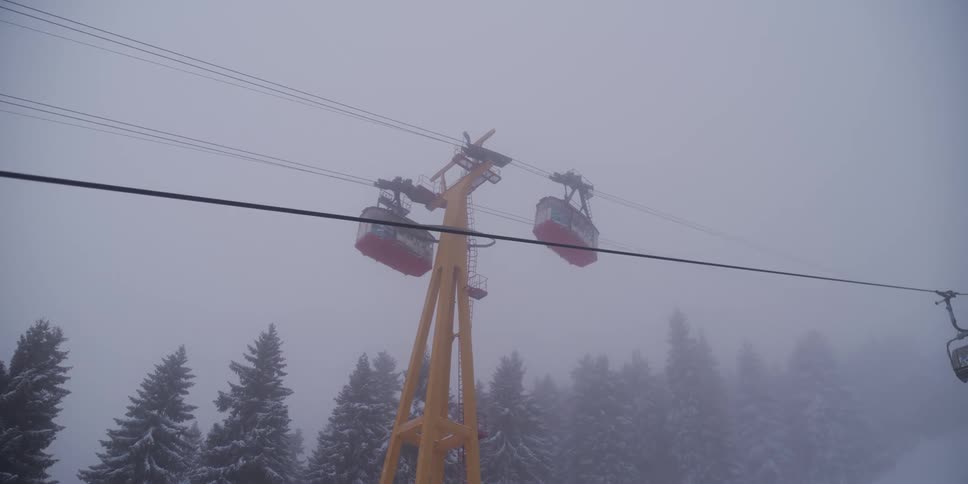A slow-motion view from a ski lift shows red gondolas moving past a yellow tower through thick fog on a snowy mountain