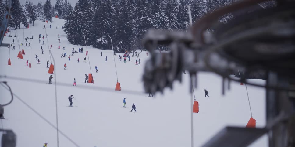 A wide shot from a ski lift shows dozens of skiers and snowboarders enjoying a busy snowy slope on a cold winter day