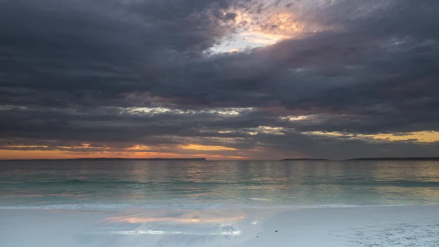 Time-lapse captures a dramatic sunrise over the ocean as waves gently wash up on a white sand beach