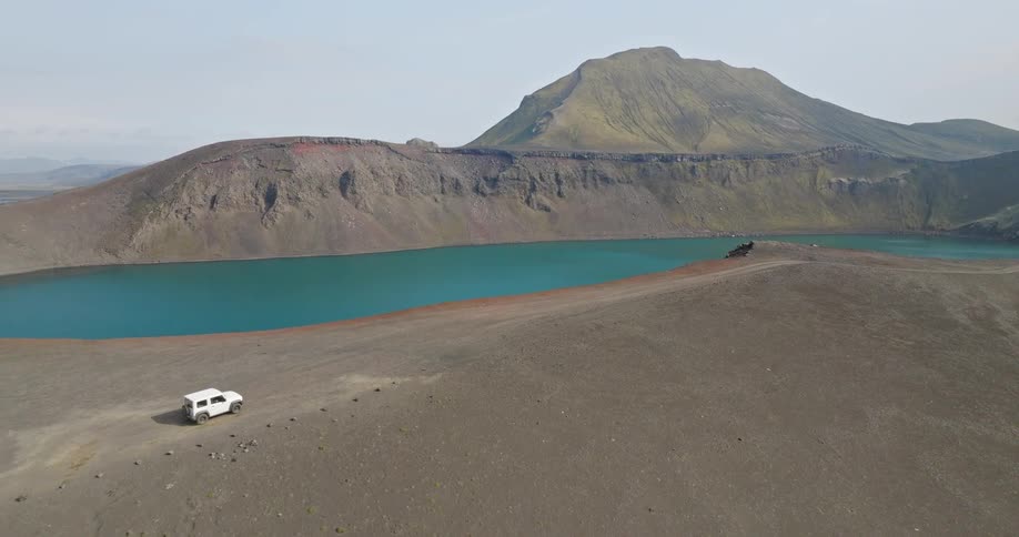 An aerial shot reveals a stunning turquoise crater lake as a white SUV drives along the volcanic rim in the Icelandic highlands
