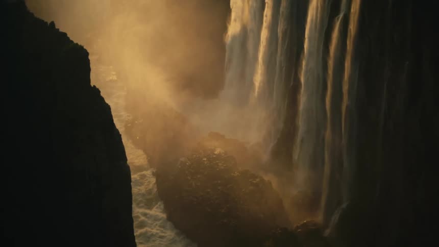 The majestic Victoria Falls plunges into a deep gorge, illuminated by the warm, golden light of sunset creating a heavy mist