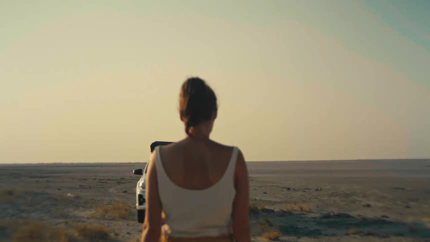 From behind, a woman walks through a vast desert landscape as her overlanding truck drives away in slow motion