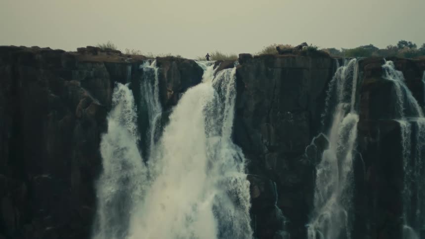 A lone figure stands atop a majestic, powerful waterfall cascading over a vast cliff