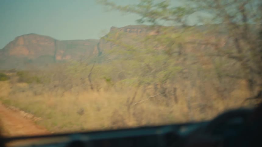 A first-person view from a safari vehicle as it follows another jeep down a dusty track through the savanna towards distant mountains
