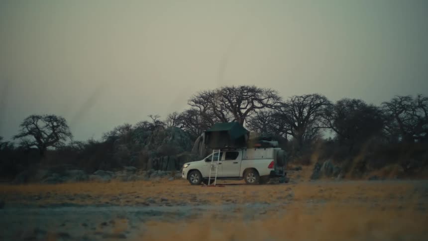 A white safari truck with a rooftop tent is parked in a remote, arid African landscape under a gloomy, overcast sky