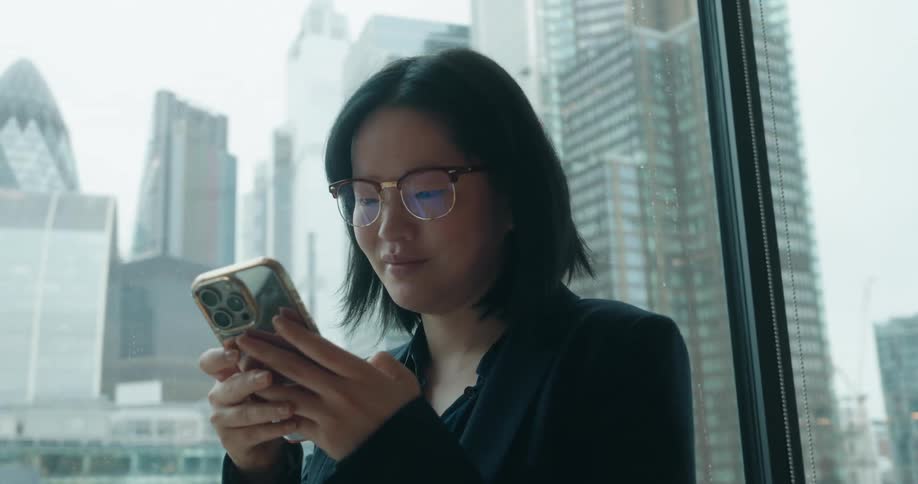 A young Asian businesswoman in glasses smiles while using her smartphone by a window overlooking the London city skyline