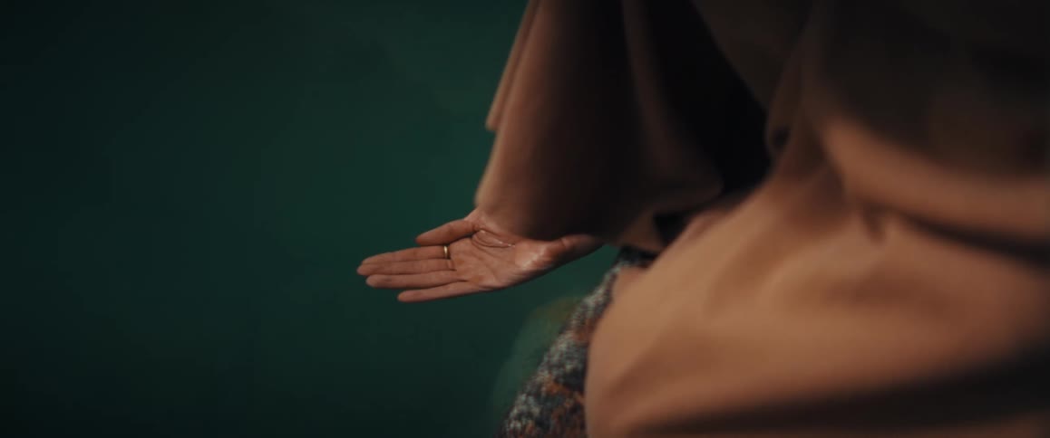 A person in a brown robe cups clear water in their hand from a dark green lake in slow motion