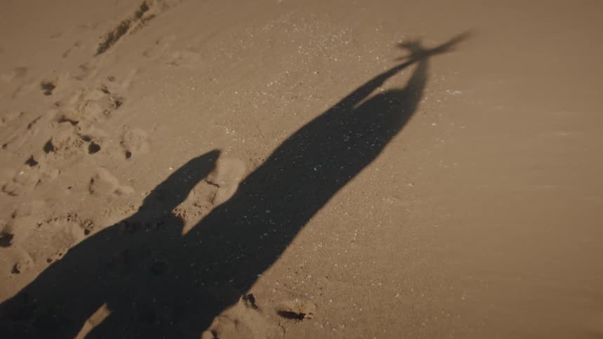 The shadow of a playful person is cast on the sand as they stretch their arms and then fly a toy airplane