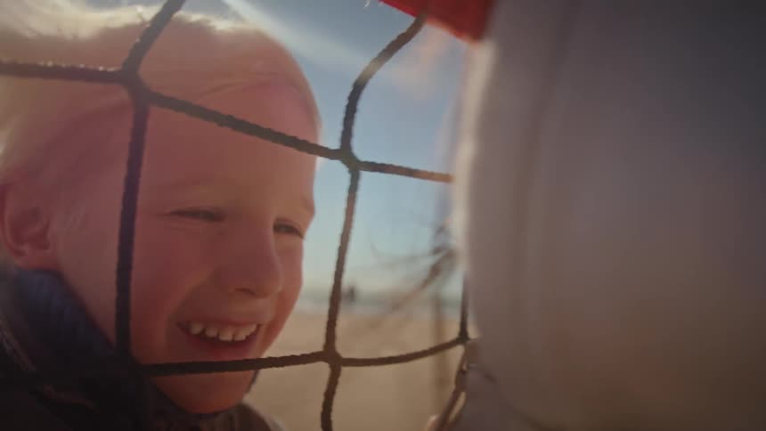 A heartwarming close-up captures a smiling young boy and his mother sharing a loving moment through a net on a windy, sunny day at the beach