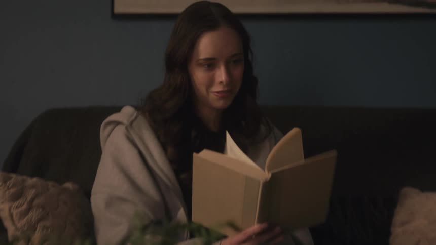 A happy young woman smiles while reading an enjoyable book on a cozy couch in a dimly lit living room at night