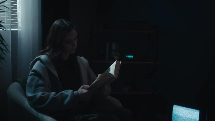 A young woman sits in a dark, moody room reading a book by the eerie glow of a static-filled television