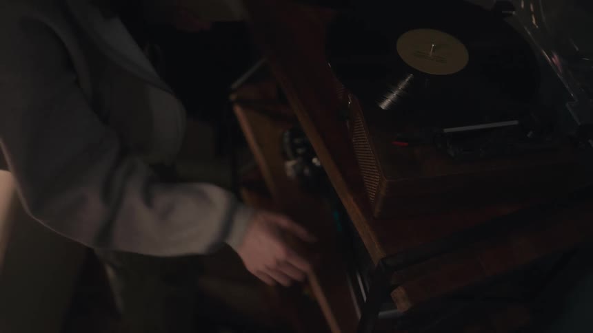 A person carefully places the needle on a spinning vinyl record playing on a vintage turntable in a cozy room