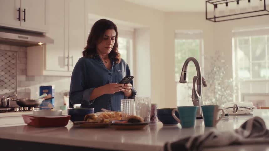 A tired woman walks into her messy kitchen and, feeling overwhelmed by the dirty dishes, pulls out her smartphone and begins to smile