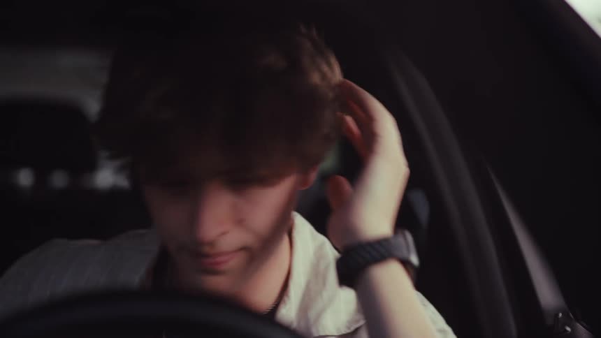 A distressed young man sits alone in his car, overcome with emotion, until he looks at his phone and his expression changes