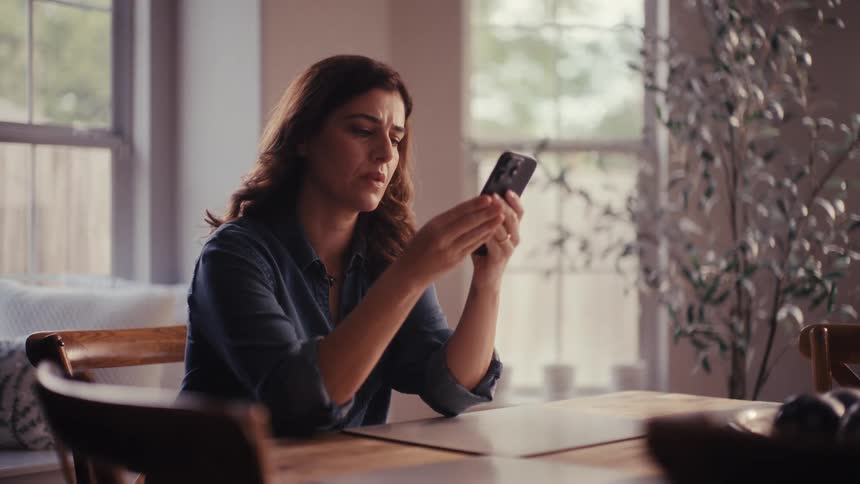 A sad and worried woman sits alone at her dining table and has an emotional reaction while looking at her phone