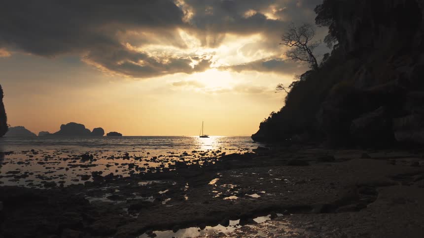 A lone sailboat is silhouetted on the ocean against a dramatic golden sunset with sunbeams breaking through the clouds