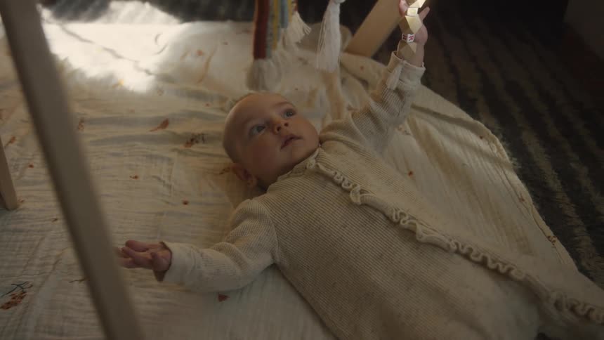 A curious baby lies on a play mat, reaching for the toys on their wooden play gym in slow motion