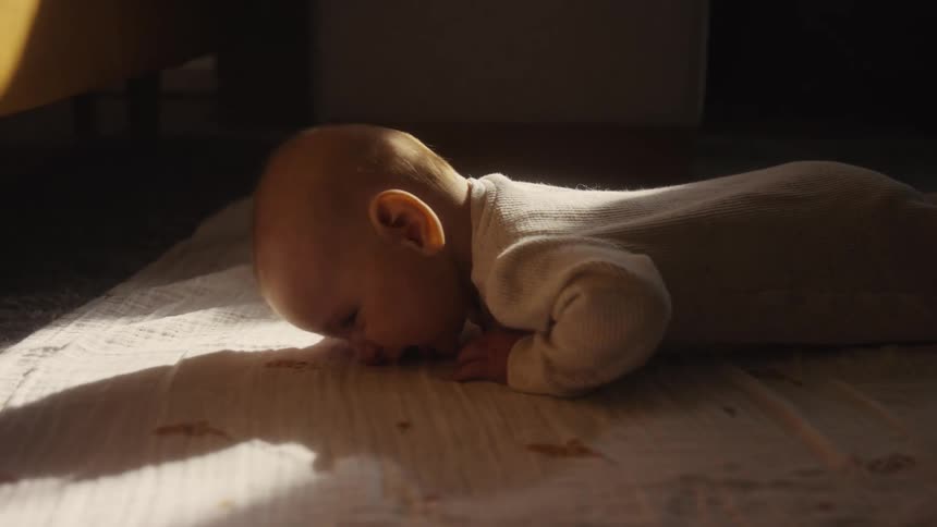 A newborn baby wearing a white onesie cries while lying on a soft blanket in a beam of warm sunlight