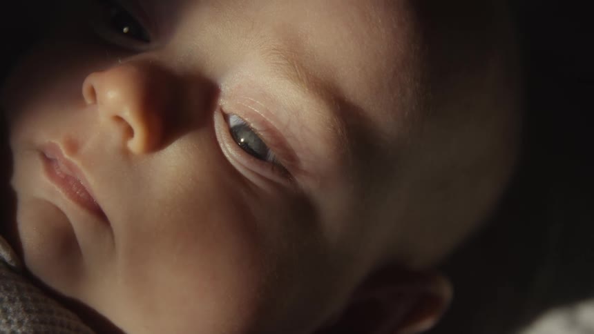 A dramatic close-up of a sleepy newborn baby's face with their eyes slowly closing in a dark room