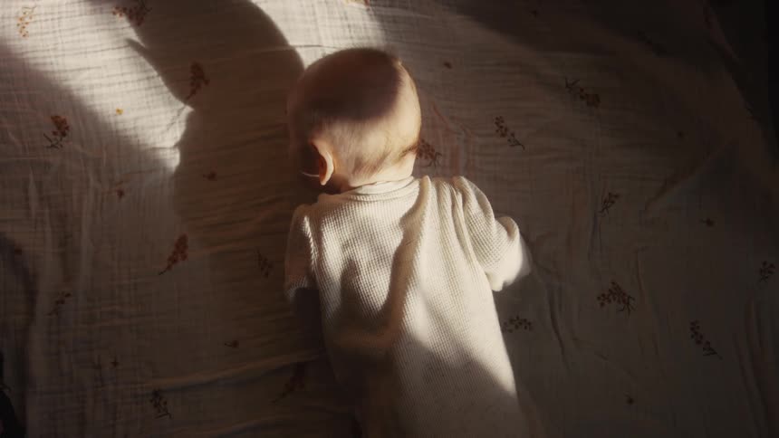 A peaceful baby sleeps on its stomach in a warm patch of sunlight, seen from a top-down perspective
