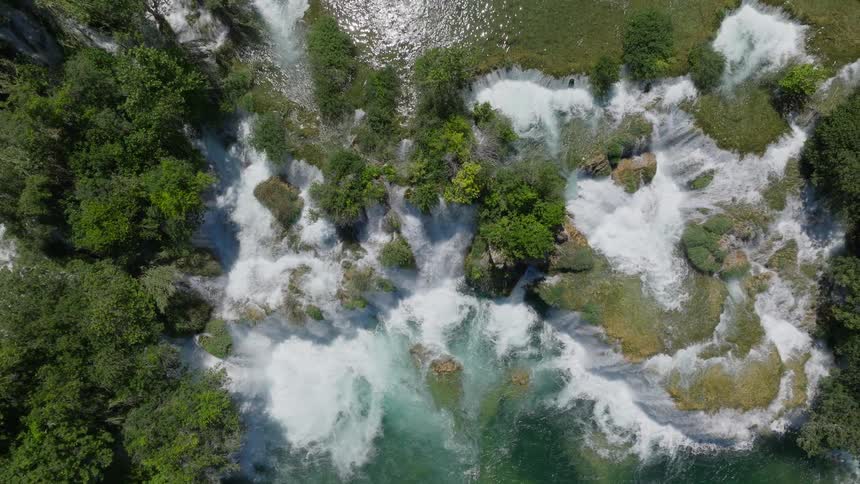 A breathtaking top-down aerial view pulls up from a powerful waterfall to reveal a stunning cascade system in a lush, green forest