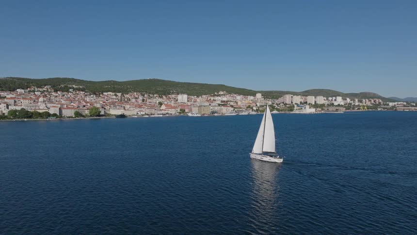 Aerial view of a white sailboat sailing across the calm blue Adriatic Sea towards the historic coastal city of Sibenik, Croatia