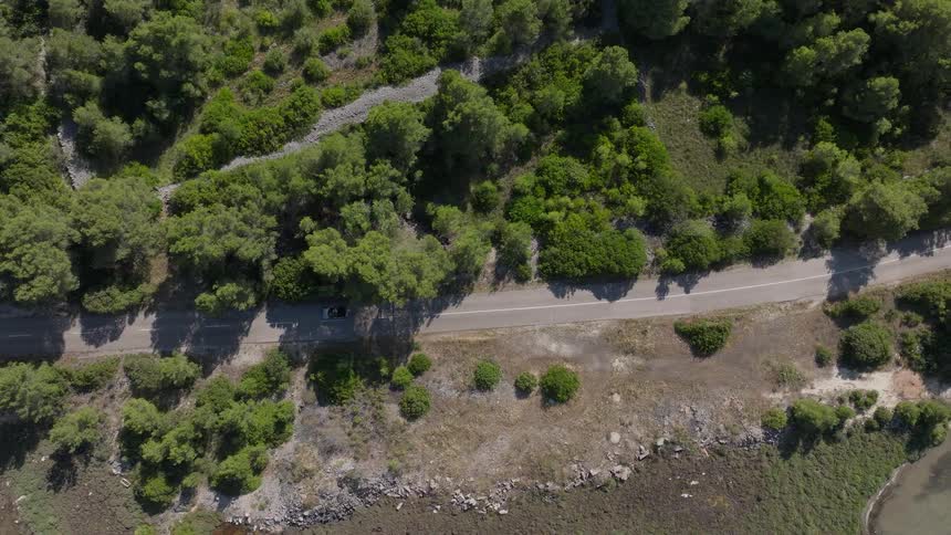 A top-down aerial view shows a lone convertible driving along a coastal road surrounded by a vibrant green Mediterranean landscape