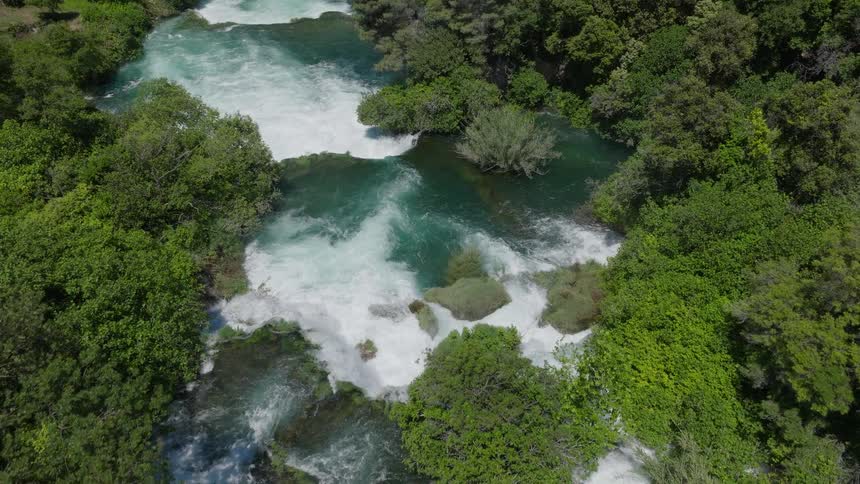 A breathtaking aerial flight over a lush green canyon reveals the stunning turquoise waterfalls of the Krka River, tilting down for a dramatic top-down view
