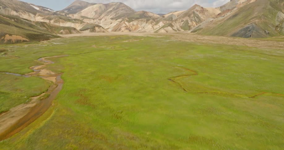 An aerial drone shot reveals a lush green valley with a winding river and colorful rhyolite mountains in the Icelandic Highlands