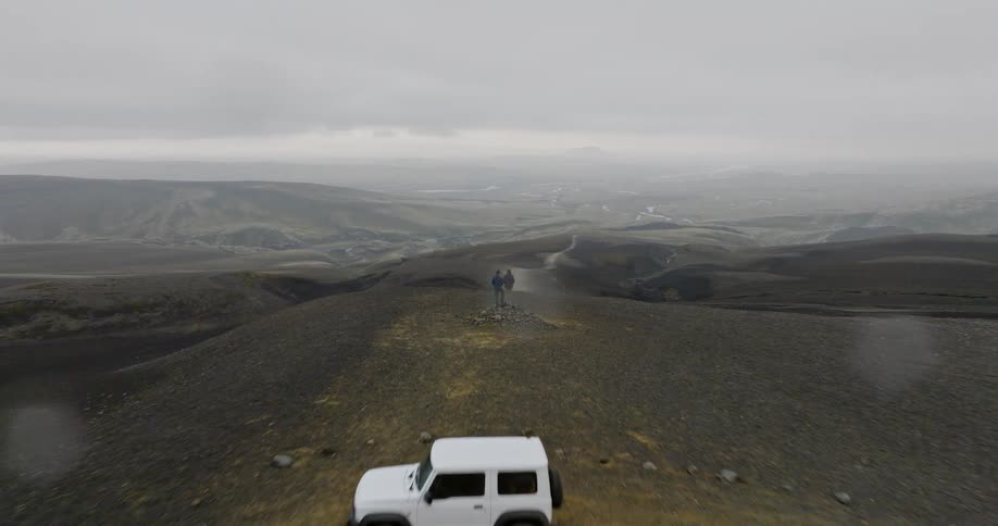 An aerial pull-back shot reveals a couple standing on a foggy, volcanic hilltop next to their 4x4, overlooking the vast and desolate landscape