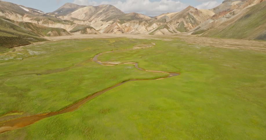 Aerial view reveals a vibrant green valley with a meandering river, set against the backdrop of the colorful Landmannalaugar mountains in the Icelandic Highlands