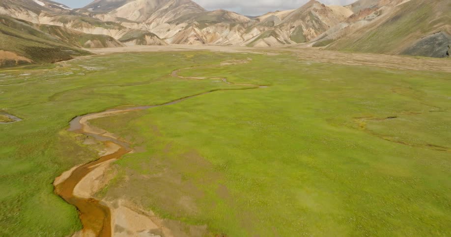 Aerial view pulls back over a meandering river in a lush green valley to reveal the colorful rhyolite mountains of Landmannalaugar, Iceland
