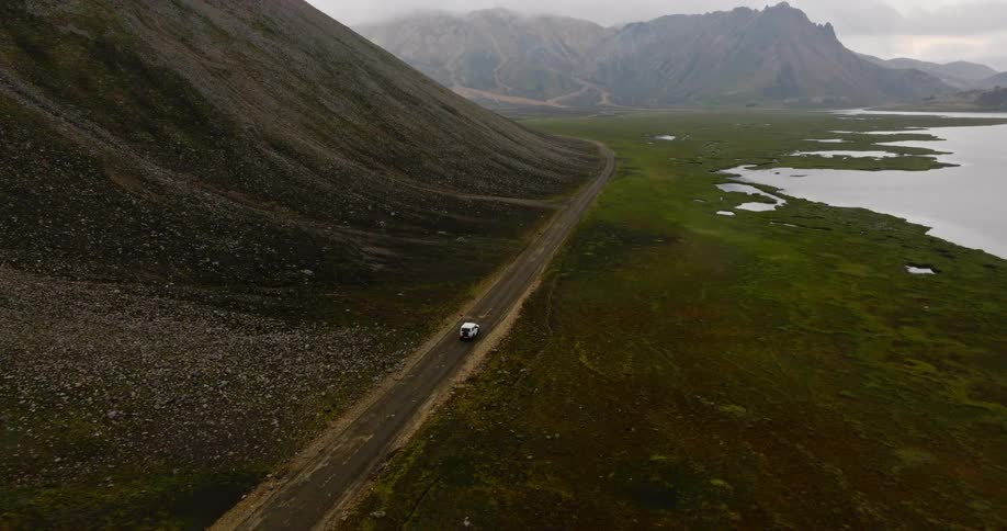An aerial view follows a lone white van driving down a remote dirt road through a vast valley in the moody Icelandic highlands