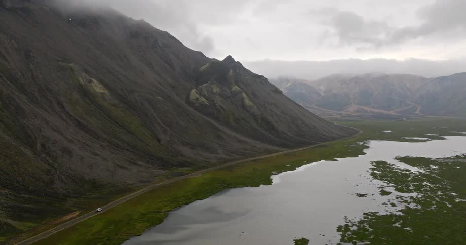 Aerial view of a solitary car driving along a remote road between a massive mountain and a tranquil lake in an epic, cloudy landscape
