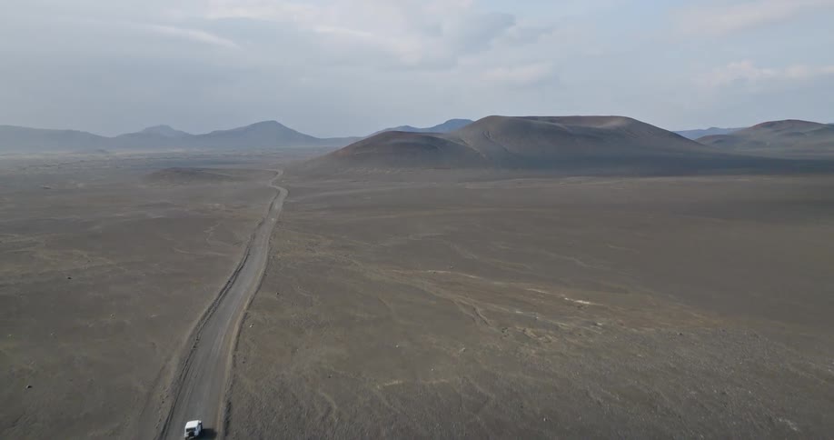 An aerial shot follows a lone white van driving down a dusty road through a vast, desolate volcanic desert