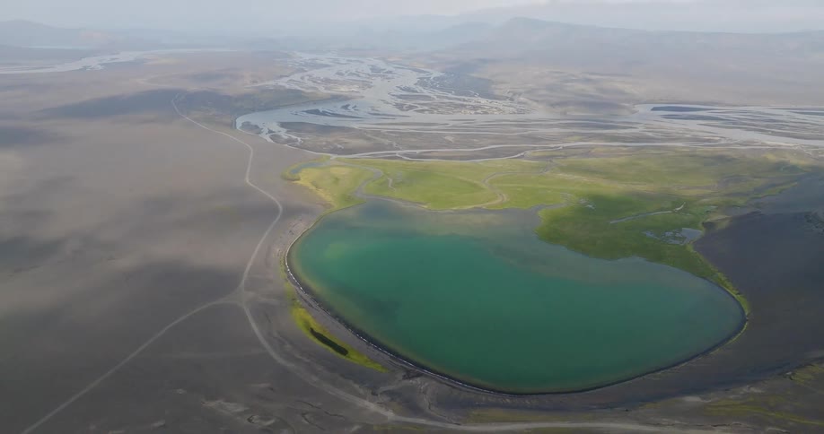 High aerial view tilts up to reveal a turquoise lake and braided river system in a desolate black sand volcanic landscape