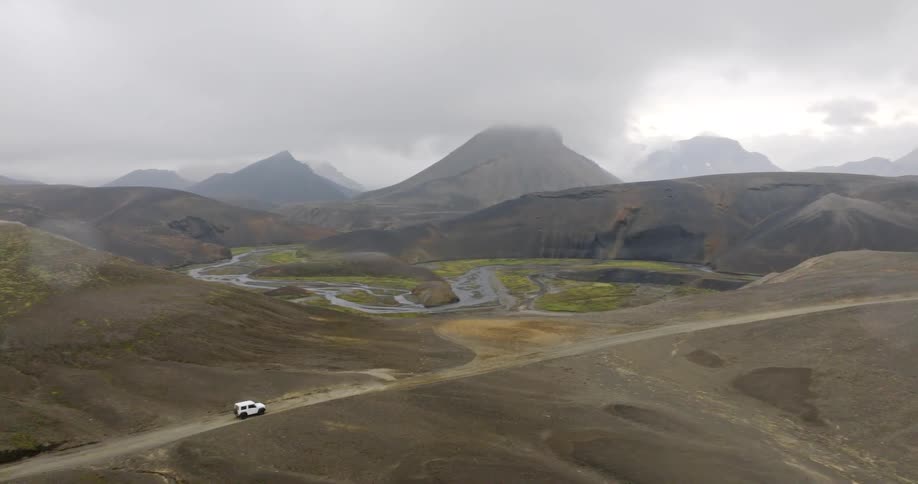 An epic aerial shot reveals a white 4x4 driving along a remote dirt road through the vast and desolate volcanic highlands of Iceland on a foggy day