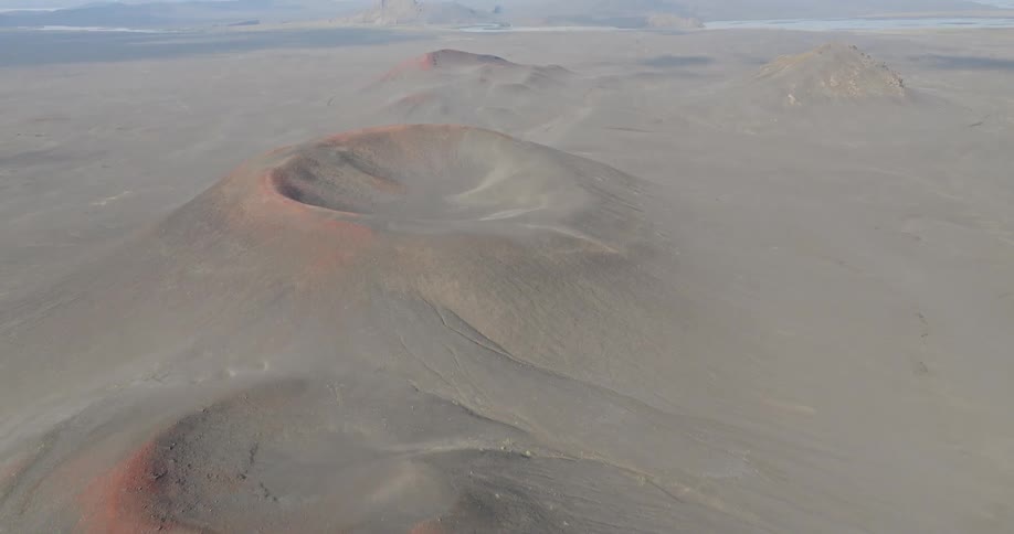 An aerial pullback shot reveals a vast and desolate volcanic landscape with several red and black craters