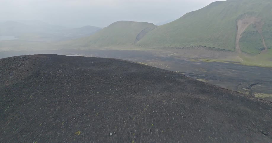An epic aerial reveals two hikers walking over a black volcanic hill, showcasing the vast, misty landscape of Iceland's highlands