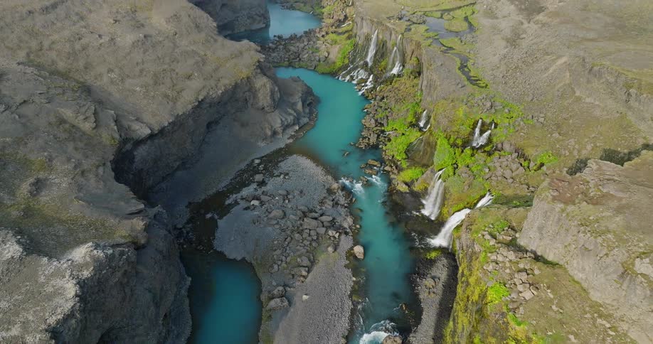 An epic aerial view reveals a hidden canyon oasis with a turquoise river and a series of cascading waterfalls in a barren Icelandic landscape