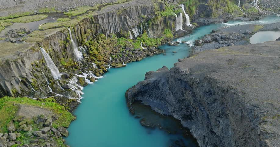 An aerial view flies over a desolate, rocky landscape to reveal a magnificent canyon with a turquoise river and numerous waterfalls cascading down the mossy cliffs