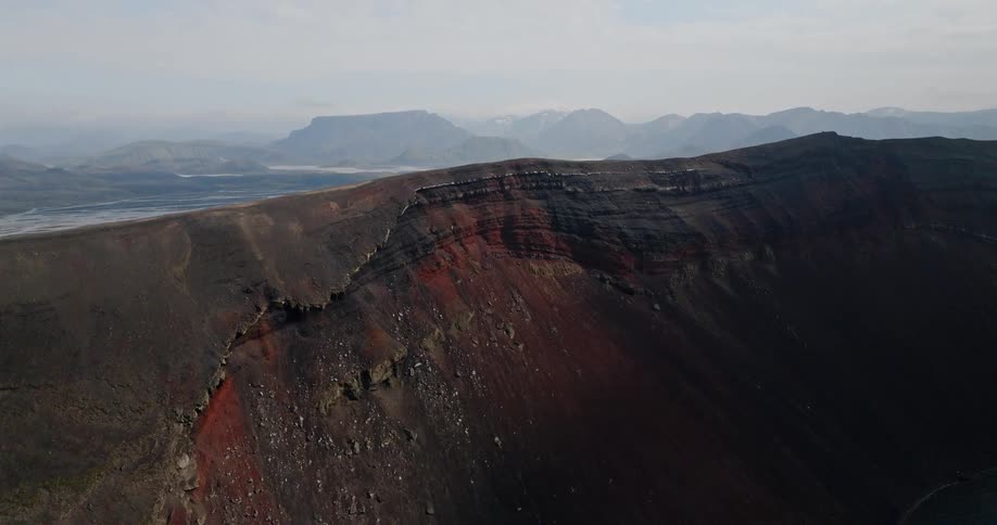 Dramatic aerial drone shot ascends over a red and black volcanic crater to reveal a vast, misty mountain landscape in Iceland