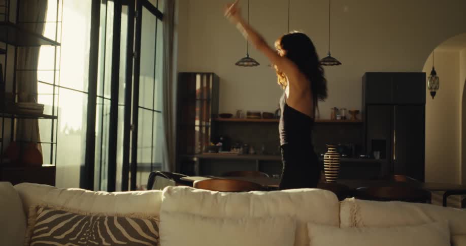 A carefree young woman in a stylish black dress dances joyfully and energetically alone in her sunlit modern home