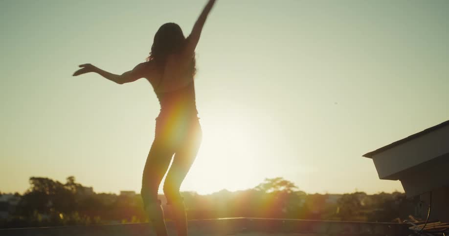 A silhouetted woman dances freely and joyfully on a rooftop as the golden sun sets behind her in slow motion